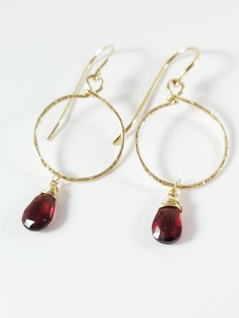 Puede incluir: Aretes de aro de oro con una piedra preciosa roja colgante. Los aros son texturizados y las piedras preciosas son facetadas.