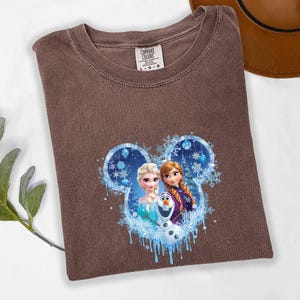 May include: A brown t-shirt with a graphic of Elsa, Anna, and Olaf from the film Frozen, set within a Mickey Mouse ear outline with snowflakes and blue accents. The shirt has a crew neck and a Comfort Colors tag.