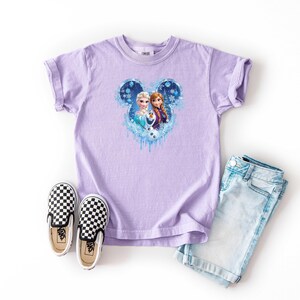 May include: A lilac t-shirt with a Frozen graphic of Elsa and Anna in a Mickey Mouse shape. The outfit includes light blue denim shorts and black and white chequered slip-on shoes.