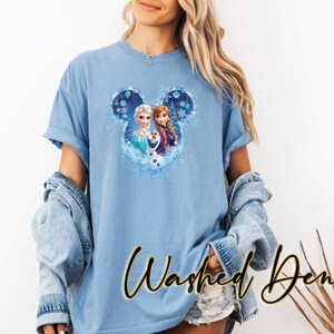 May include: Pale blue t-shirt with a Frozen graphic of Elsa, Anna, and Olaf inside a Mickey Mouse ear shape. The sleeves are rolled up, and the words "Washed Denim" are written in a script font.