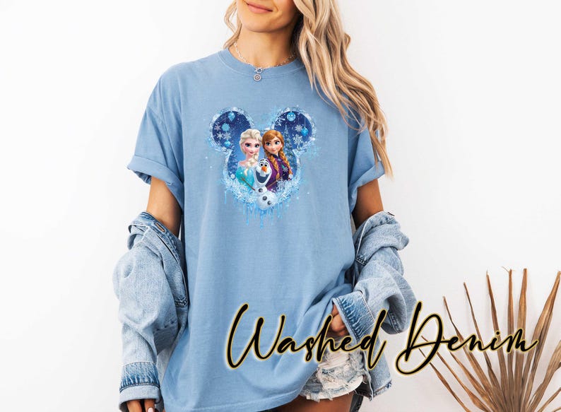May include: Pale blue t-shirt with a Frozen graphic of Elsa, Anna, and Olaf inside a Mickey Mouse ear shape. The sleeves are rolled up, and the words "Washed Denim" are written in a script font.