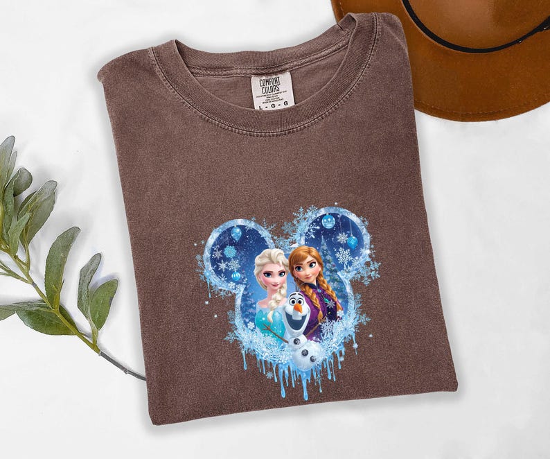 May include: A brown t-shirt with a graphic of Elsa, Anna, and Olaf from the film Frozen, set within a Mickey Mouse ear outline with snowflakes and blue accents. The shirt has a crew neck and a Comfort Colors tag.