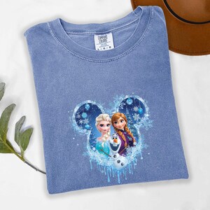 May include: A light blue Comfort Colors t-shirt with a graphic of Elsa, Anna, and Olaf from Frozen within a Mickey Mouse-shaped design. The design includes snowflakes and icy accents. The shirt is laid flat on a white surface.