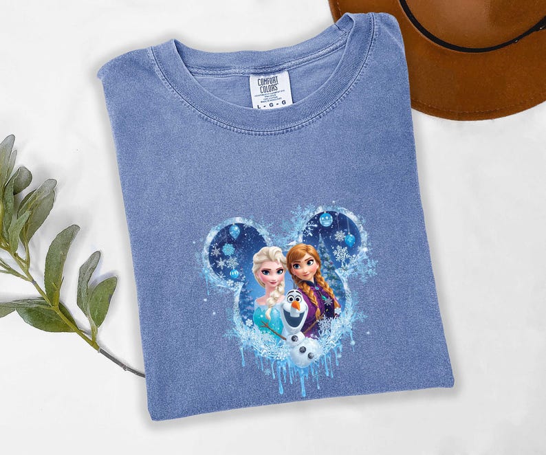 May include: A light blue Comfort Colors t-shirt with a graphic of Elsa, Anna, and Olaf from Frozen within a Mickey Mouse-shaped design. The design includes snowflakes and icy accents. The shirt is laid flat on a white surface.