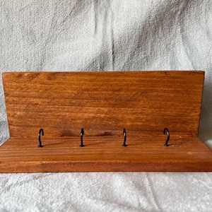 Può includere: Un portachiavi in legno tinto, di colore marrone rossiccio caldo, con quattro ganci in metallo nero. Il portachiavi ha una forma rettangolare con un retro inclinato e una base piatta. I ganci sono distanziati uniformemente.