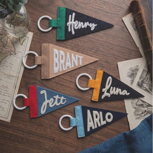 May include: Five triangular felt pennant keychains with white plastic rings. The pennants feature names in white lettering: Henry on dark blue, BRANT on tan, Jett on blue with a red header, Luna on black with a yellow header, and ARLO on dark blue with a light blue header.