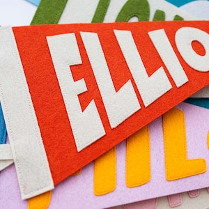Peut inclure: Gros plan sur des fanions en feutre colorés. Le fanion principal est orange avec le nom "ELLIO" en lettres blanches. D'autres fanions sont visibles en bleu, vert et rose, avec d'autres noms en jaune et rose.