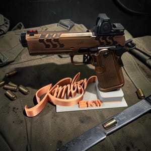 May include: A bronze-colored handgun with a red dot sight, resting on a gray and white stand. The stand features the name "Kimber" in a 3D-printed orange script. Several bullets and a knife are also visible.