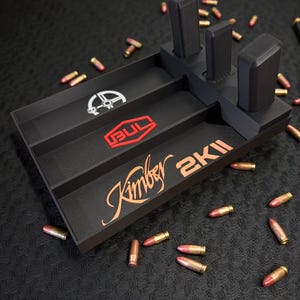 Puede incluir: Organizador negro con el texto "Kimber 2K11" en escritura color cobre. El organizador tiene tres ranuras verticales y dos compartimentos con el logotipo "BUL" y un gráfico blanco. Varias balas están esparcidas alrededor del organizador.