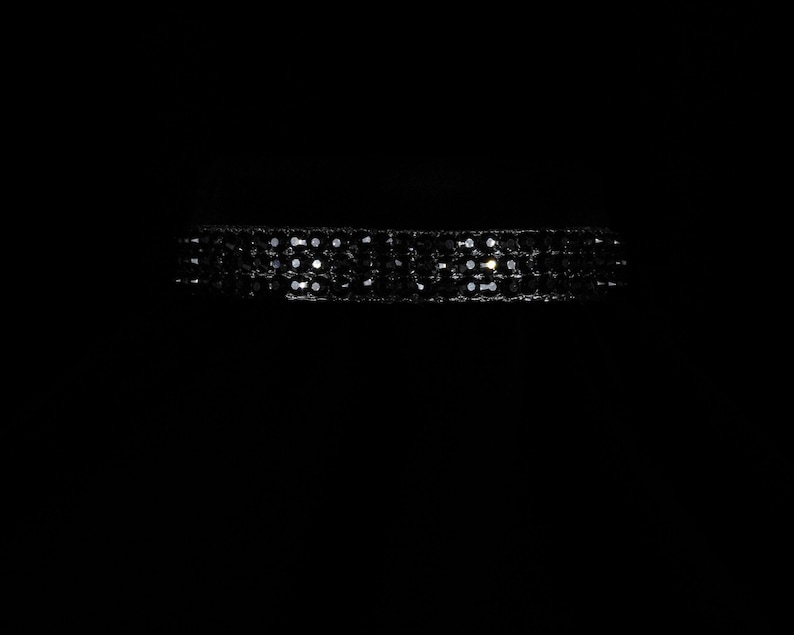 K&ouml;nnte beinhalten: Ein schwarzes Choker-Halsband mit mehreren Reihen kleiner, funkelnder Verzierungen. Das Halsband ist vor einem dunklen Hintergrund platziert, was die reflektierende Qualit&auml;t der Verzierungen hervorhebt. Das Design deutet auf ein formelles oder Abendaccessoire hin.