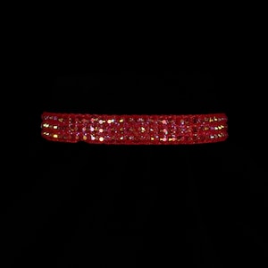May include: A red, sparkly bracelet against a black background. The bracelet is a band of red fabric covered in small, glittering sequins. The sequins catch the light, creating a shimmering effect. The bracelet appears to be a choker or wristband.