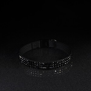 K&ouml;nnte beinhalten: Ein schwarzes Choker-Halsband mit Reihen kleiner, funkelnder schwarzer Edelsteine. Das Choker hat ein schwarzes Stoffband und einen Schnallenverschluss. Der Schmuck wird auf einer dunklen, reflektierenden Oberfl&auml;che pr&auml;sentiert.