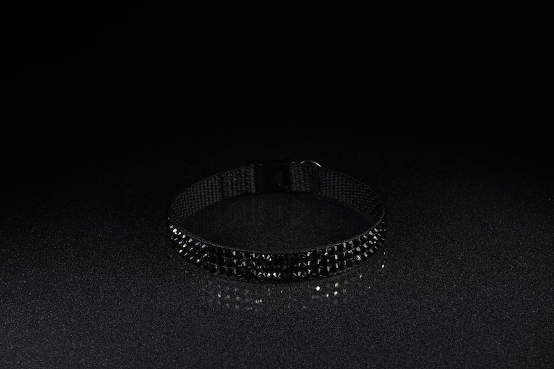 K&ouml;nnte beinhalten: Ein schwarzes Choker-Halsband mit Reihen kleiner, funkelnder schwarzer Edelsteine. Das Choker hat ein schwarzes Stoffband und einen Schnallenverschluss. Der Schmuck wird auf einer dunklen, reflektierenden Oberfl&auml;che pr&auml;sentiert.