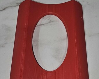 May include: A red, 3D-printed plastic stand featuring a curved, rectangular base with a large, centred oval cutout. The object has a smooth, matte finish and visible vertical layer lines from the manufacturing process, with a concave top edge.