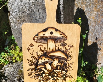 May include: A wooden cutting board with a handle features a design of mushrooms burned into the surface. The mushrooms are brown and white with black spots. The design includes leaves and flowers.
