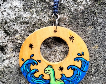 May include: A circular wooden pendant featuring a hand-painted illustration of a green sea creature emerging from blue ocean waves. The design includes small black star accents and is suspended from a dark blue cord by a silver metal jump ring.
