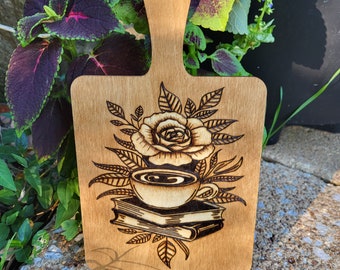 May include: A wooden cutting board with a hand-burned design of a coffee cup, a rose, and books. The design is in shades of brown and black.