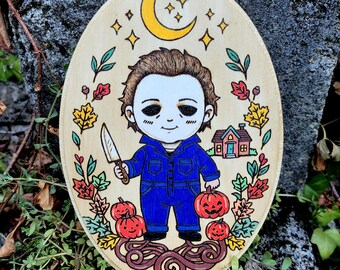 May include: An oval wooden plaque featuring a wood-burned and colored illustration of a character with a white mask, blue jumpsuit, and knife. The design includes a yellow crescent moon, stars, autumn leaves, three jack-o'-lanterns, and a house.