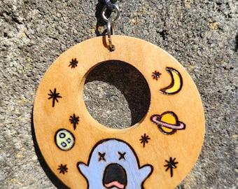 May include: A circular wooden pendant featuring a hand-painted light blue ghost with an open mouth and x-shaped eyes, surrounded by a yellow crescent moon, a yellow planet with a pink ring, and several small black star symbols.