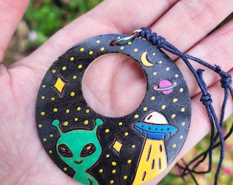May include: A circular black pendant featuring a hand-painted space scene with a green alien, a blue and yellow UFO, a pink planet, a yellow crescent moon, and yellow stars. The pendant is attached to a dark blue cord with adjustable knots.