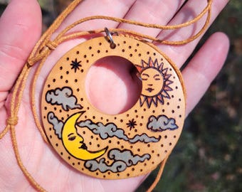 May include: A circular wooden pendant on a tan cord featuring a wood-burned design of a yellow crescent moon, a sun with a face, and grey clouds. The surface is decorated with small dots and star shapes, with a central circular cutout.