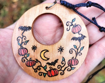 May include: A circular wooden pendant featuring a wood-burned design of orange pumpkins, black bats, a yellow crescent moon, and stars. The pendant has a central circular cutout and is attached to a thin black cord through a metal jump ring.