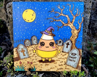 May include: A Halloween-themed wood-burned art piece featuring a cute candy corn character wearing a witch hat, standing in a graveyard under a full moon. The artwork is on a square wooden panel with a blue starry sky background.