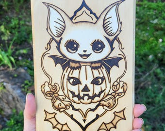 May include: A wood-burned image of a white bat with large eyes sitting on top of a jack-o'-lantern. The bat has wings and a small mouth. The jack-o'-lantern has a carved face with a wide smile and two eyes.