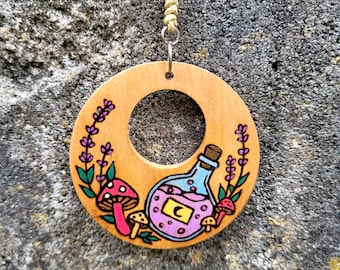 May include: A circular wooden pendant hanging from a tan cord. The wood features a hand-painted design of a light blue potion bottle with a yellow crescent moon label, pink liquid, purple lavender sprigs, and red and brown mushrooms.