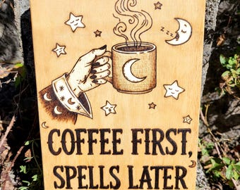 May include: A rectangular wooden plaque featuring a pyrography design of a hand holding a steaming mug with a crescent moon icon. The text reads COFFEE FIRST, SPELLS LATER, surrounded by various star and moon illustrations on the light wood surface.