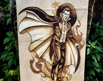 May include: Wood-burned artwork of a vampire in a black cape and suit, with a stern expression. The artwork is on a light-colored wooden board, with the image created using a pyrography technique. The vampire is surrounded by stylized flames.