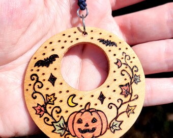May include: A circular wooden pendant featuring a wood-burned design of a jack-o'-lantern, autumn leaves, bats, a crescent moon, and stars. The light-colored wood ornament is suspended from a dark blue cord by a silver-toned metal jump ring.