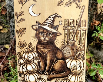 May include: A rectangular wooden plaque featuring a pyrography illustration of a cat wearing a pointed hat. The cat sits among pumpkins and oak leaves, with a crescent moon and a broomstick leaning against a wooden fence in the background.