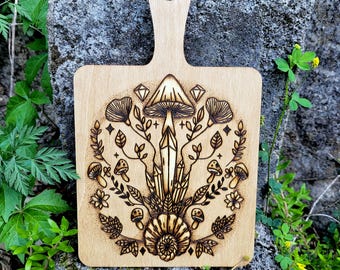 May include: A light-colored wooden cutting board with a handle. The board features a pyrography design of a mushroom, crystals, and botanical elements. The design is centered on the board and surrounded by a handle with a hole.