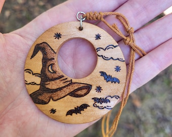 May include: A circular light-colored wooden ornament featuring a wood-burned design of a pointed hat, clouds, stars, and bats. The piece includes a silver metal jump ring attached to a brown knotted cord for hanging purposes.