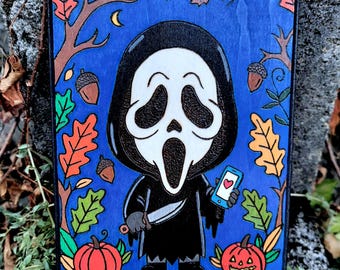 May include: A rectangular wooden plaque featuring a cartoon-style figure in a black hooded robe and white mask, holding a knife and a blue smartphone with a heart icon. The background is blue with colorful autumn leaves, acorns, and two pumpkins.
