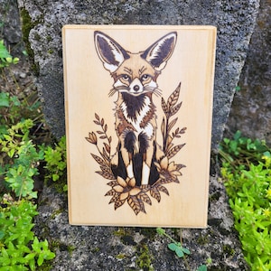 May include: A wood burned design of a fennec fox sitting in a bed of flowers. The fox is brown and white with large ears. The flowers are stylized and have a whimsical feel.
