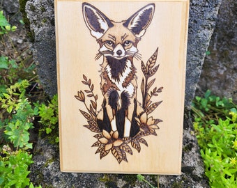 May include: A wood burned design of a fennec fox sitting in a bed of flowers. The fox is brown and white with large ears. The flowers are stylized and have a whimsical feel.