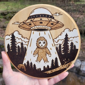 May include: A wood-burned round wooden plaque with a cartoon image of a Bigfoot being abducted by a UFO. The UFO is hovering over the Bigfoot, and there are trees in the background.