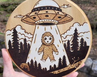 May include: A wood-burned round wooden plaque with a cartoon image of a Bigfoot being abducted by a UFO. The UFO is hovering over the Bigfoot, and there are trees in the background.