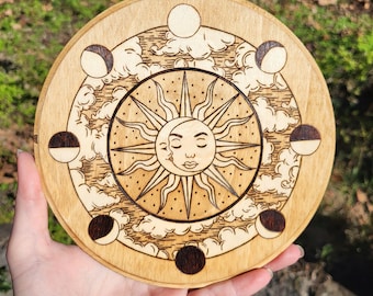 May include: A wooden round plaque with a sun and moon design. The sun has a face and is surrounded by rays. The moon is in different phases around the sun. The plaque is brown and has a natural wood finish.