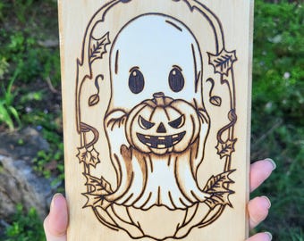 May include: A wood burned design of a ghost holding a jack-o-lantern. The ghost has large eyes and a friendly expression. The jack-o-lantern has a toothy grin.