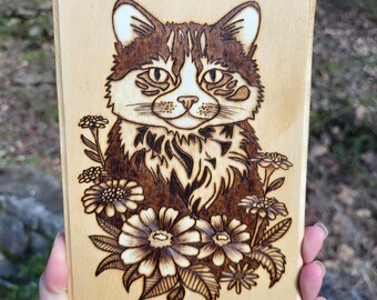 May include: A wood-burned image of a cat with brown and white fur, surrounded by flowers. The cat is looking directly at the viewer.