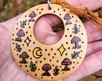 May include: A circular wooden pendant featuring wood-burned illustrations of mushrooms, small leaves, a crescent moon, and four-pointed stars. The design is accented with light pink and green coloring, and the piece hangs from a brown cord.
