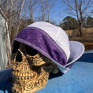 May include: A white welding cap with a purple paisley band and white stitching. The cap is displayed on a decorative skull with floral carvings. The skull is tan and sits on a blue surface.