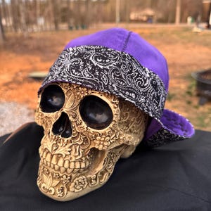 May include: A decorative skull with intricate floral carvings wears a purple and black paisley bandana. The skull is beige and rests on a black surface, with a blurred outdoor background.