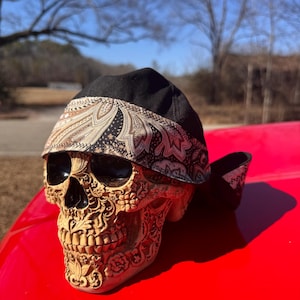 May include: A decorative skull with intricate floral carvings, topped with a black and paisley patterned head covering. The skull rests on a bright red surface, with a blurred outdoor background.