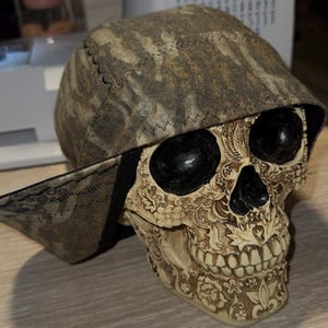 May include: A decorative skull with a camouflage-patterned cap. The skull is off-white with intricate floral and leaf carvings. The cap has a brown and tan pattern. The eyes are black and glossy.