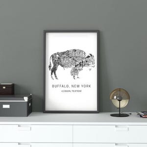 Peut inclure: Une impression encadrée en noir et blanc représentant une carte de Buffalo, New York, en forme de bison. L'impression comprend le nom et les coordonnées de la ville. L'impression est sur un meuble blanc avec des objets décoratifs.