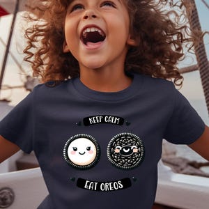 May include: Navy blue t-shirt featuring two cartoon Oreo cookies with smiling faces. The text "KEEP CALM" is above the cookies, and "EAT OREOS" is below. The shirt is worn by a child.